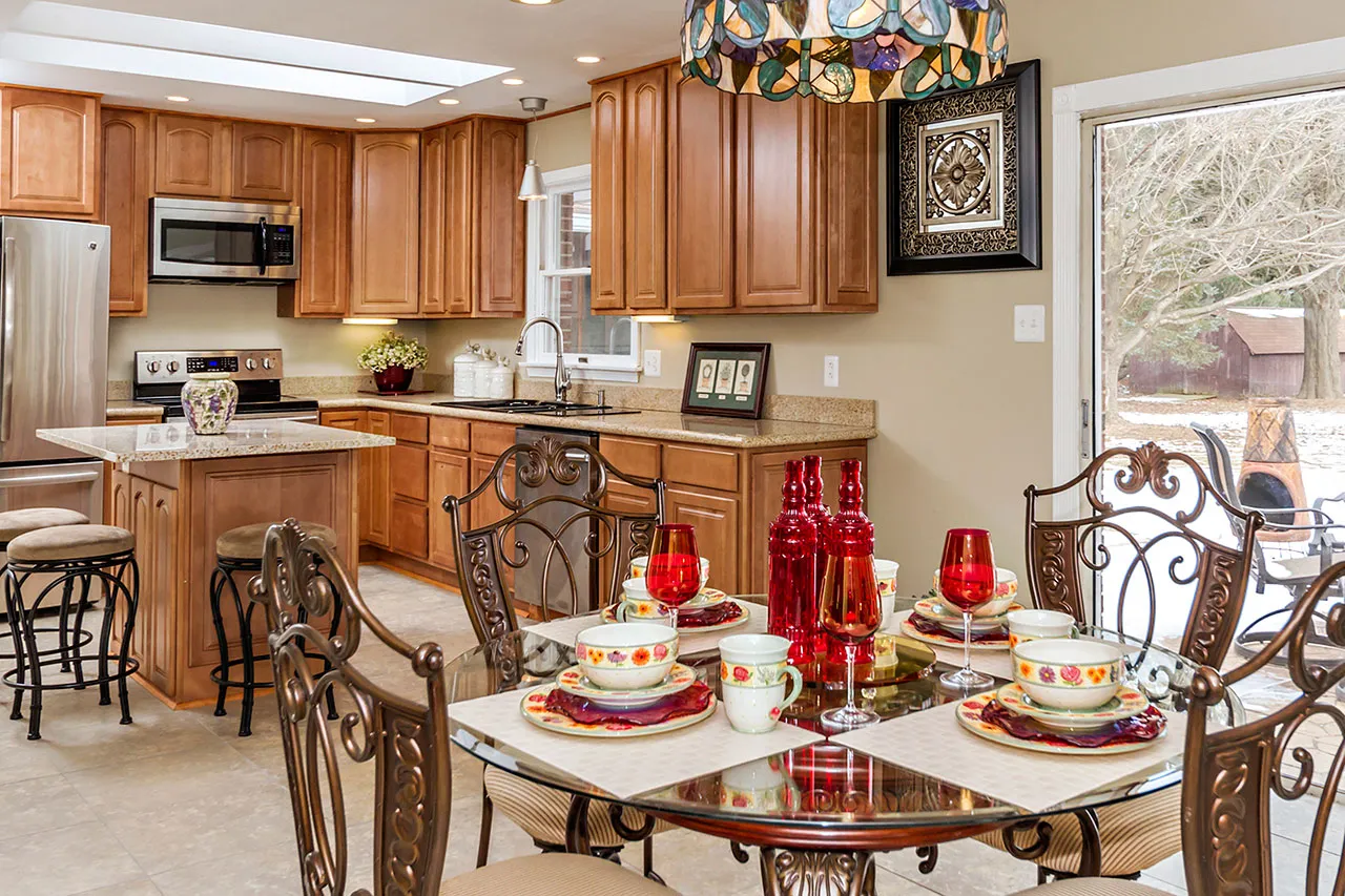 Kitchen and dining area with wooden cabinets, a glass table set with colorful dishware, red cups, decorative chandelier, and large window overlooking a snowy yard.