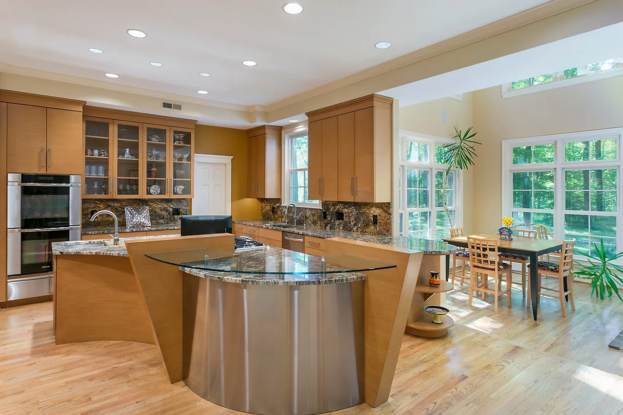 Spacious kitchen with modern wooden cabinets, stainless steel appliances, and a granite island. Adjacent dining area with large windows and plants. Natural light fills the space.