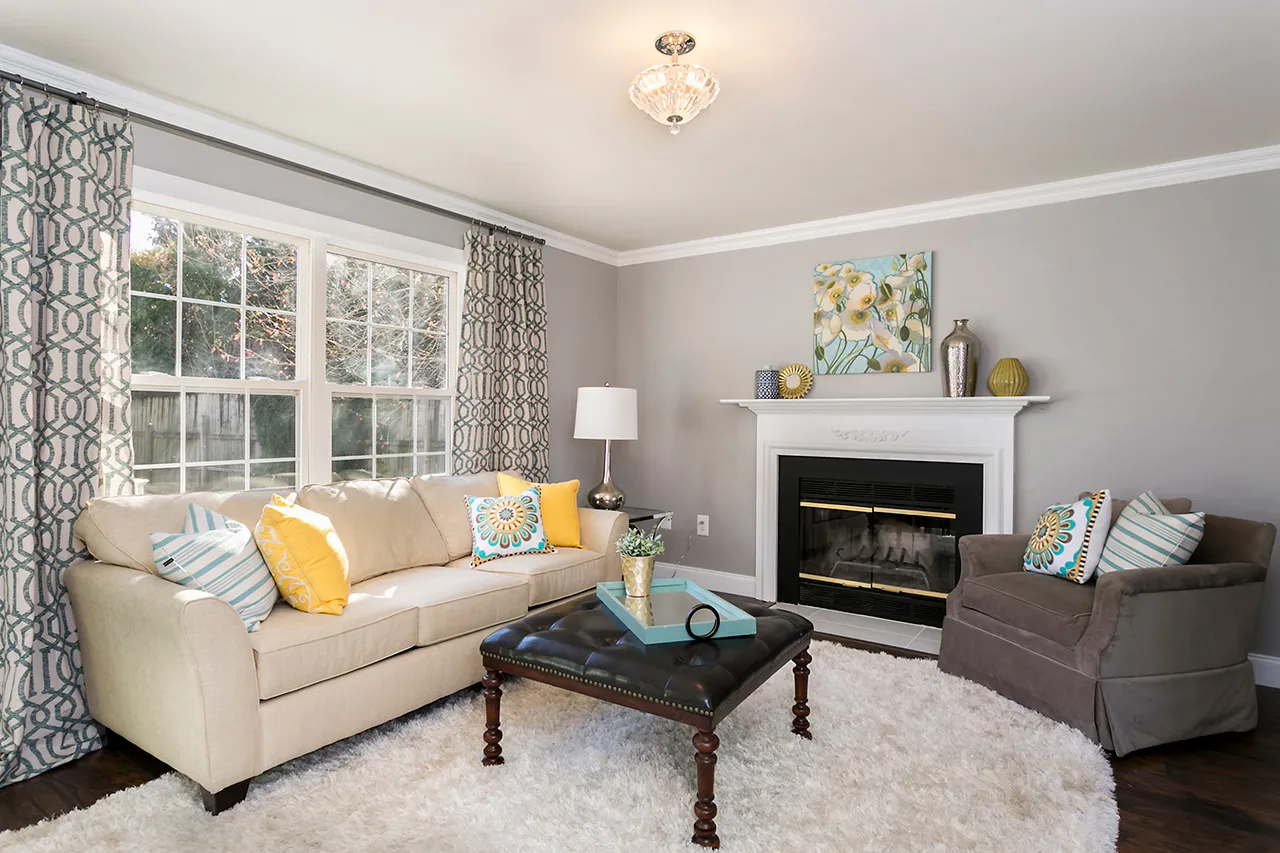 Living room with a beige sofa, gray armchair, and dark ottoman on a white rug. Decor includes a painting above the fireplace, patterned curtains, and colorful pillows.