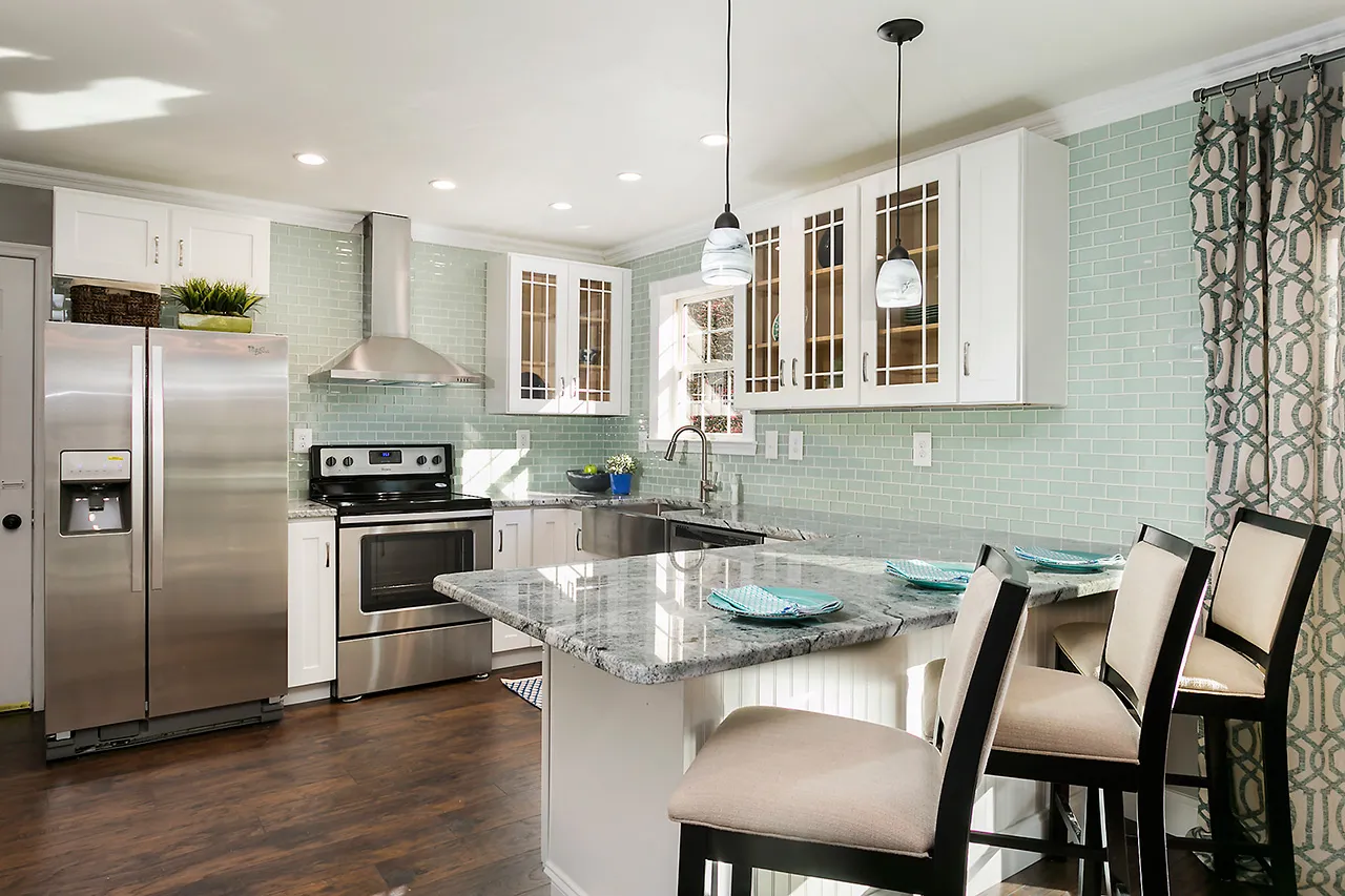 Modern kitchen with stainless steel appliances, white cabinets, teal glass tile backsplash, and a marble island with bar stools. Hanging lights illuminate the space.