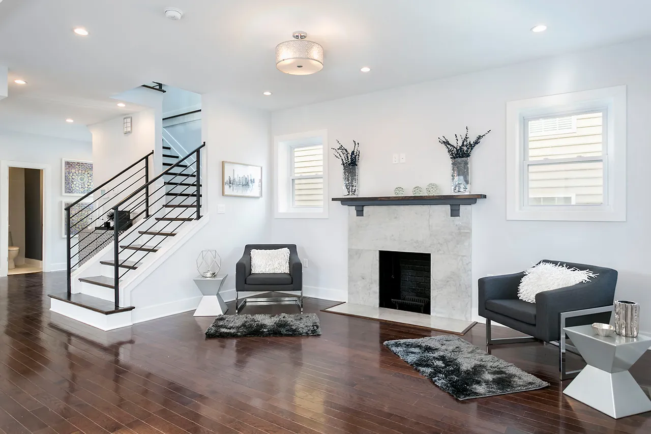 Modern living room with dark hardwood floors, a fireplace, and two gray armchairs. A metal staircase with black railings is visible. Minimalist decor includes black-and-white accents.