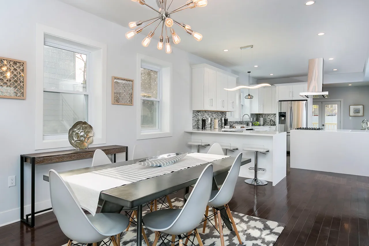 Modern dining room and kitchen with a rectangular table, white chairs, dark hardwood floors, abstract chandelier, and bar stools at a breakfast counter.