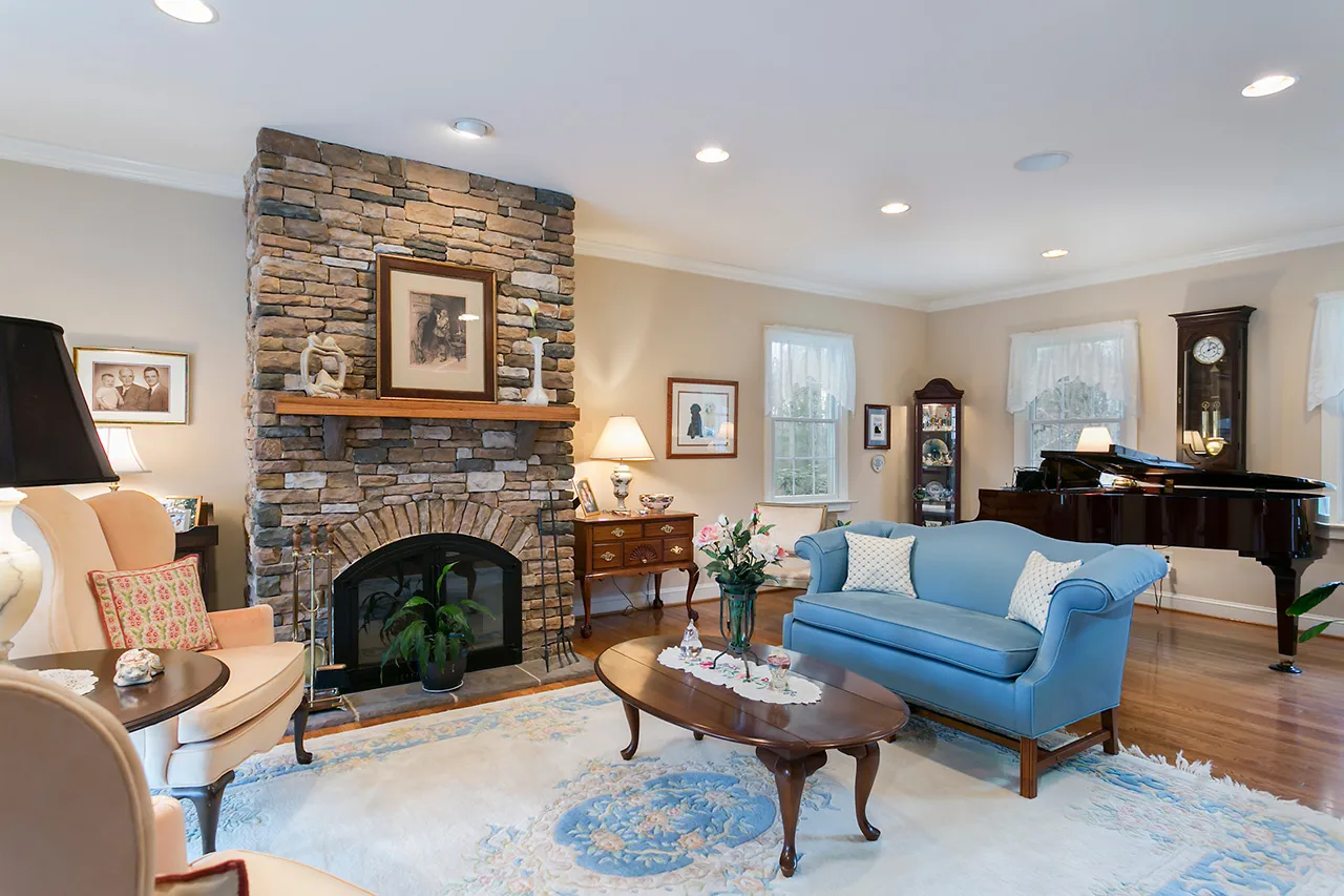 A cozy living room with a stone fireplace, blue sofa, wooden coffee table, grand piano, and framed artwork. The decor includes a grandfather clock and two armchairs.