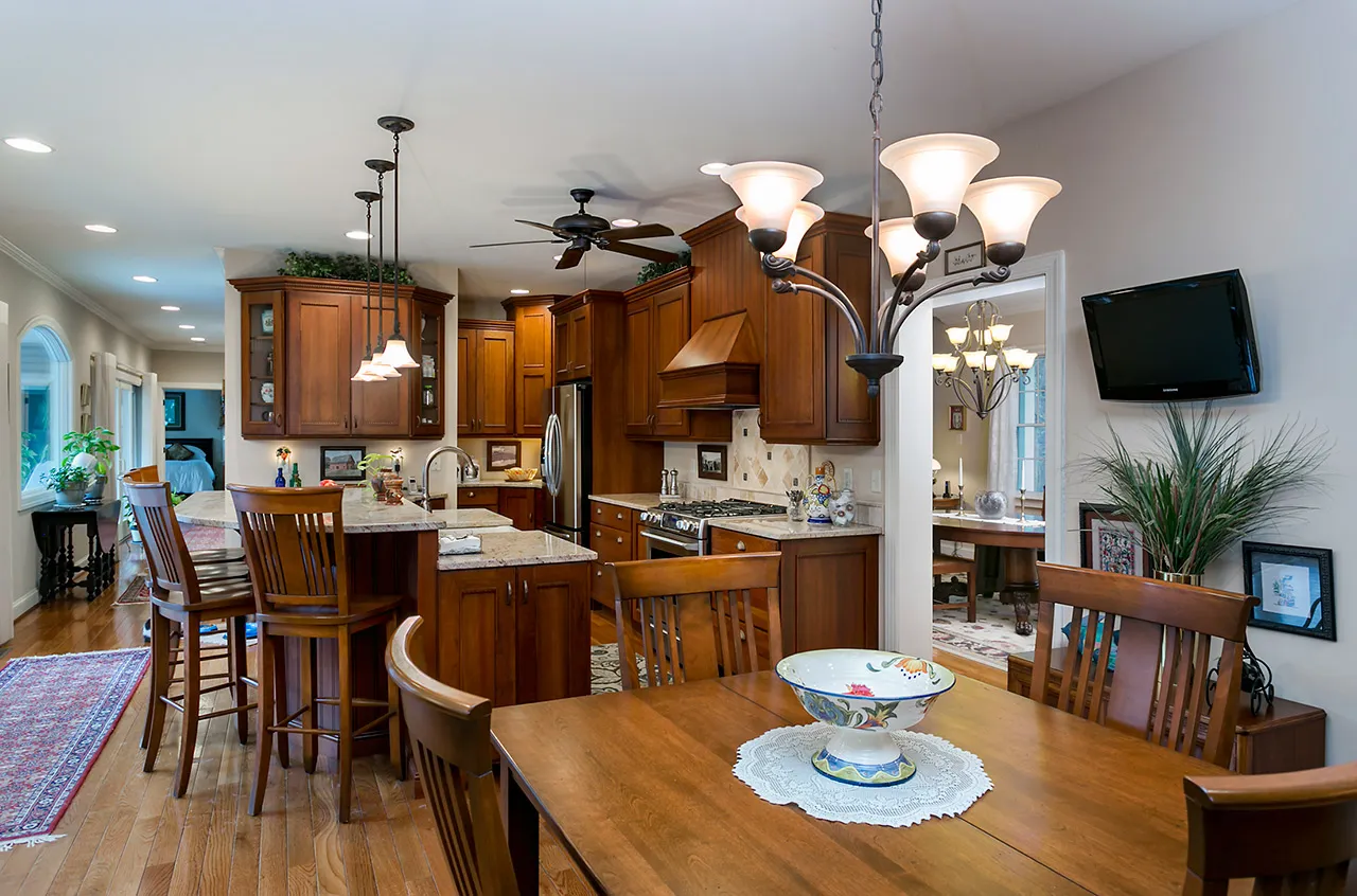 Spacious kitchen and dining area with wooden cabinets, granite countertops, and a chandelier. Includes a dining table, wooden chairs, and a wall-mounted TV.