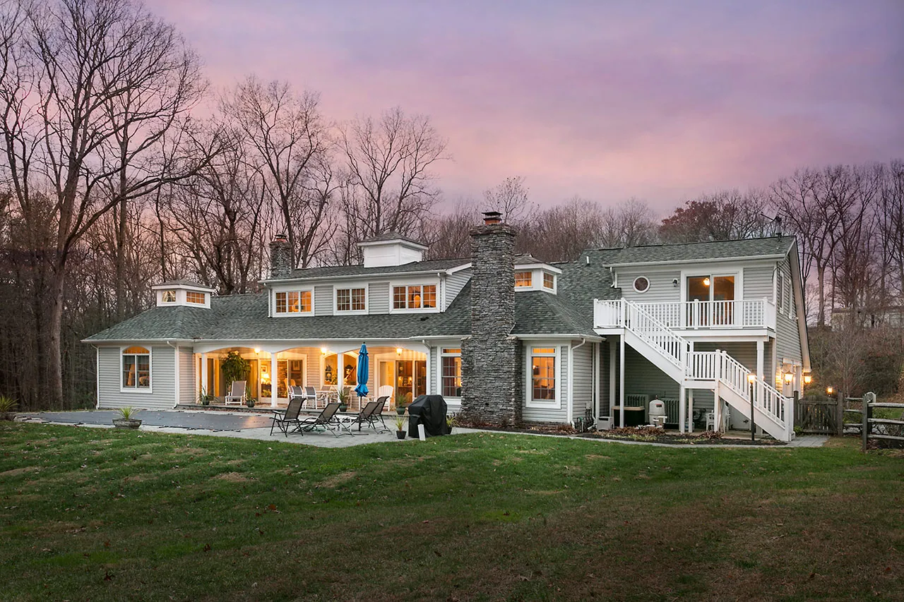 A large house with multiple windows and a stone chimney, surrounded by trees at dusk. The upper balcony has a white railing, and the backyard features a patio with outdoor furniture.