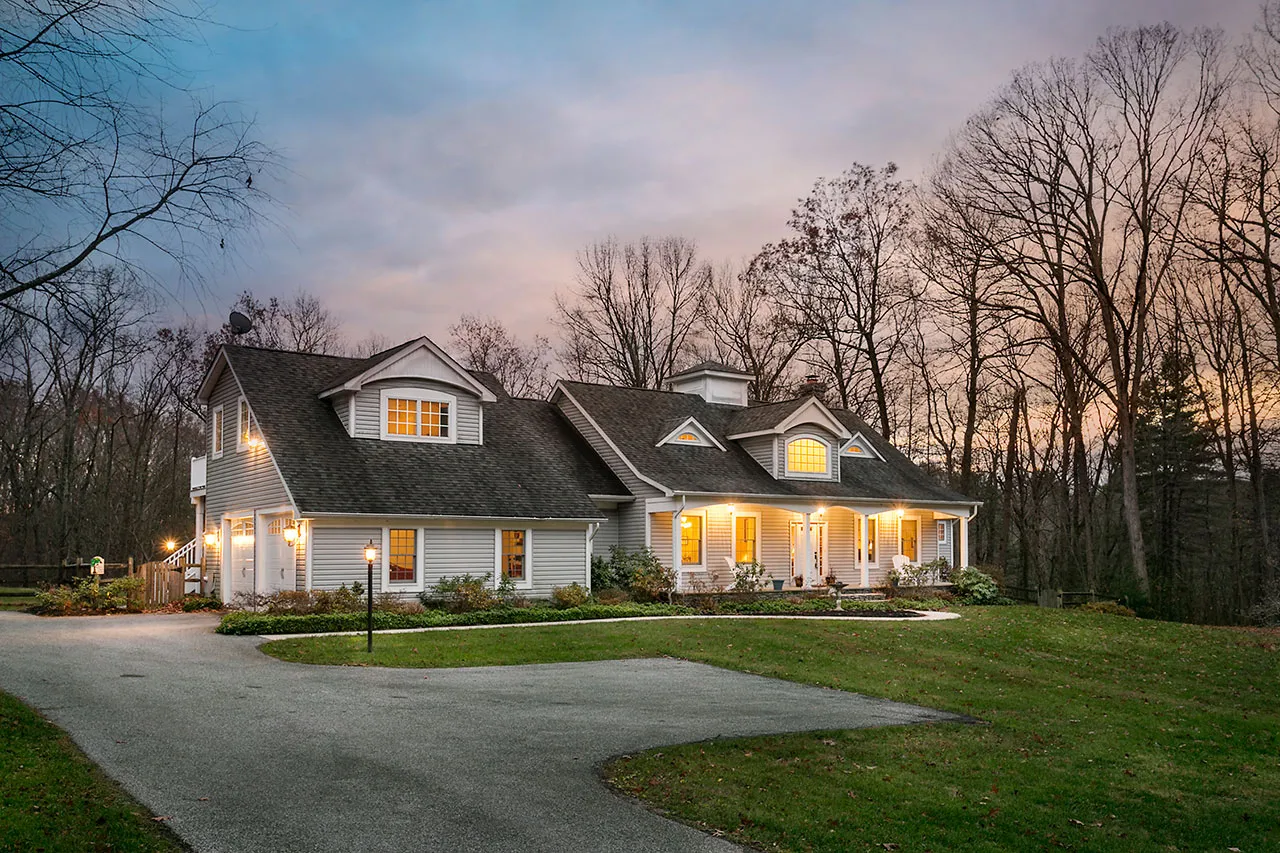 A large, two-story house with lit windows, surrounded by leafless trees, stands near a curved driveway under a dusk sky.