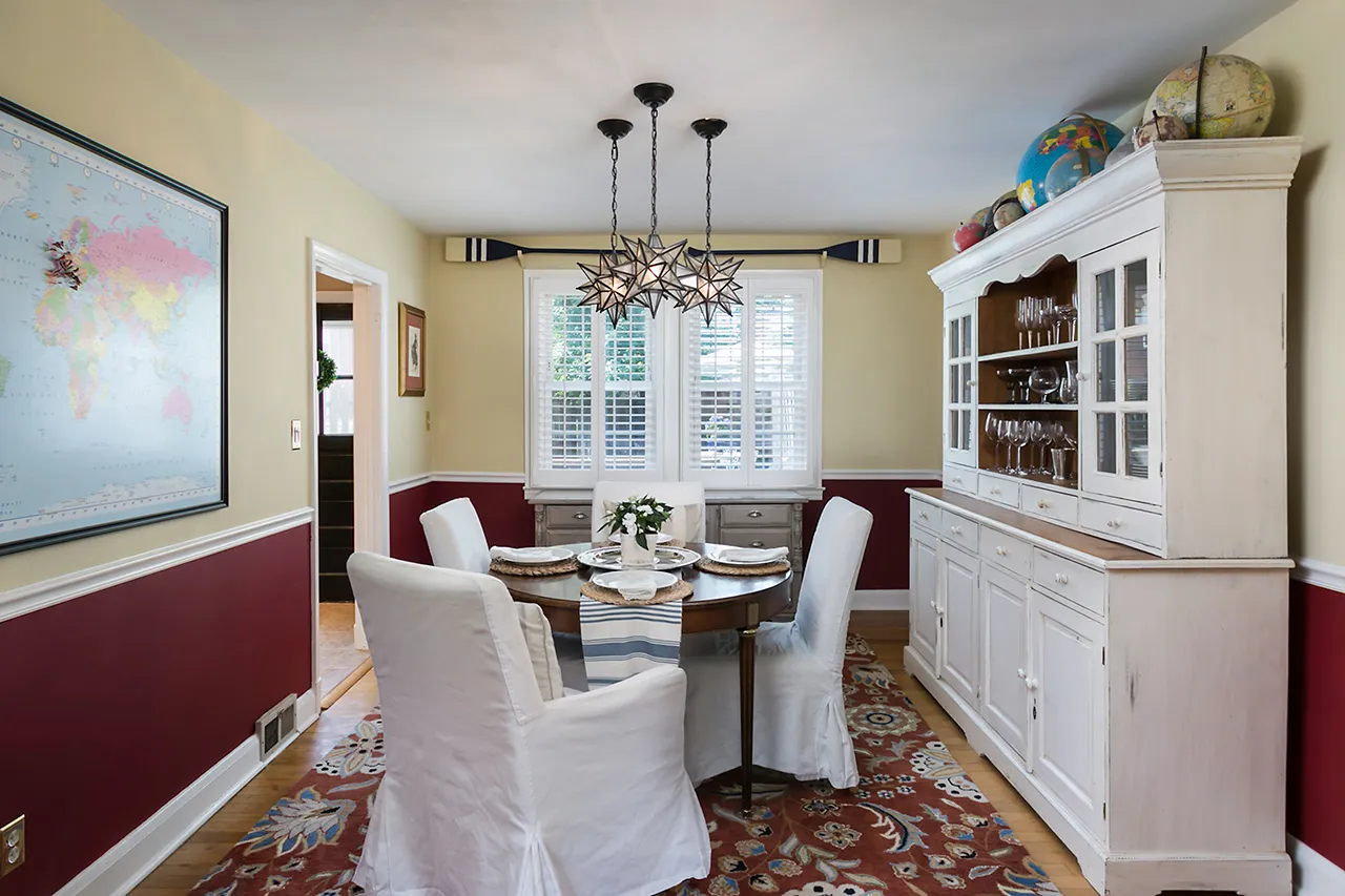 Dining room with a round table, six white slipcovered chairs, globe-topped hutch, world map on the wall, pendant lights, and a red-patterned carpet.