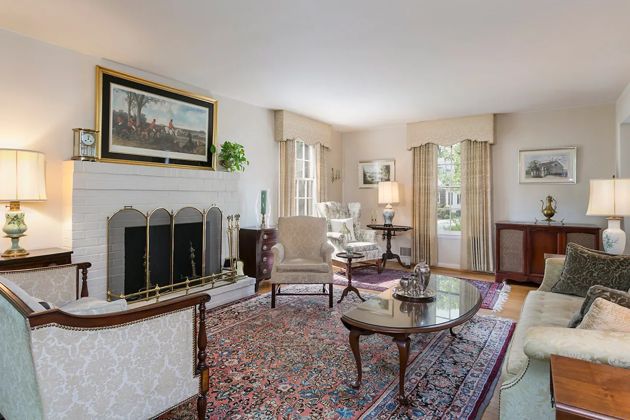 Living room with ornate rug, fireplace, seating area, coffee table, and framed artwork. Large windows allow natural light, illuminating the traditional decor.