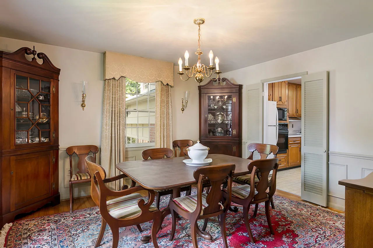 Dining room with a wooden table, six chairs, two glass cabinets, decorative vases, a chandelier, and a large rug. A doorway leads to a kitchen with wooden cabinets.