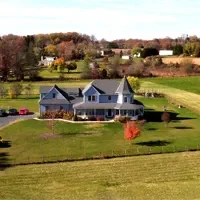 Aerial view of a large country house with a wraparound porch, surrounded by green lawns and autumn trees, set in a rural landscape with neighboring homes in the background.