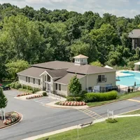 Aerial view of a building with a brown roof surrounded by trees, featuring a large rectangular swimming pool with lounge chairs on one side. A parking lot is visible in the foreground.