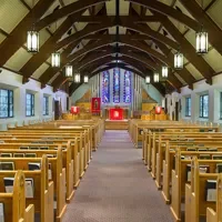 Interior of a church with wooden pews, a high vaulted ceiling, hanging lights, and a colorful stained glass window at the front.