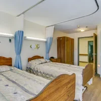 Two twin hospital beds with floral bedspreads and blue privacy curtains in a room with wooden furniture and a hallway exit.
