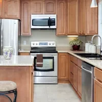 Kitchen with wooden cabinets, stainless steel appliances, including fridge, oven, and microwave. A kitchen island with a decorative vase is in the foreground. Sink and countertop on the right.