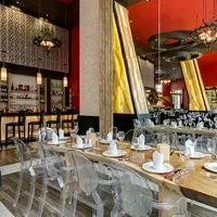 Modern restaurant interior with elegant decor, featuring a long wooden table set with white plates and glasses. Transparent chairs and gold-accented mirrors complement the red and black color scheme.