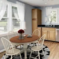 A kitchen with light wood cabinets, a round dining table with four white chairs, a vase of red flowers on the table, and two windows with white curtains.